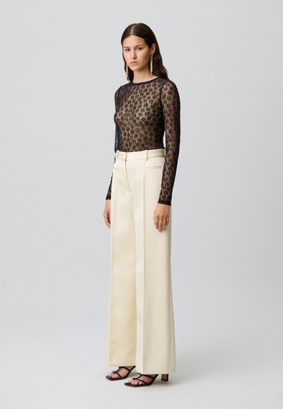 Top lungo a maniche sheer nere con un motivo floreale, abbinato a pantaloni larghi beige a vita alta. Modella che indossa tacchi neri con cinturino.