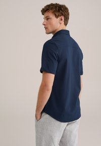 Camisa polo azul marino, de mangas cortas, con cuello plano, de textura suave, que presenta un dobladillo recto. Llevada con pantalones de cuadros gris claro.
