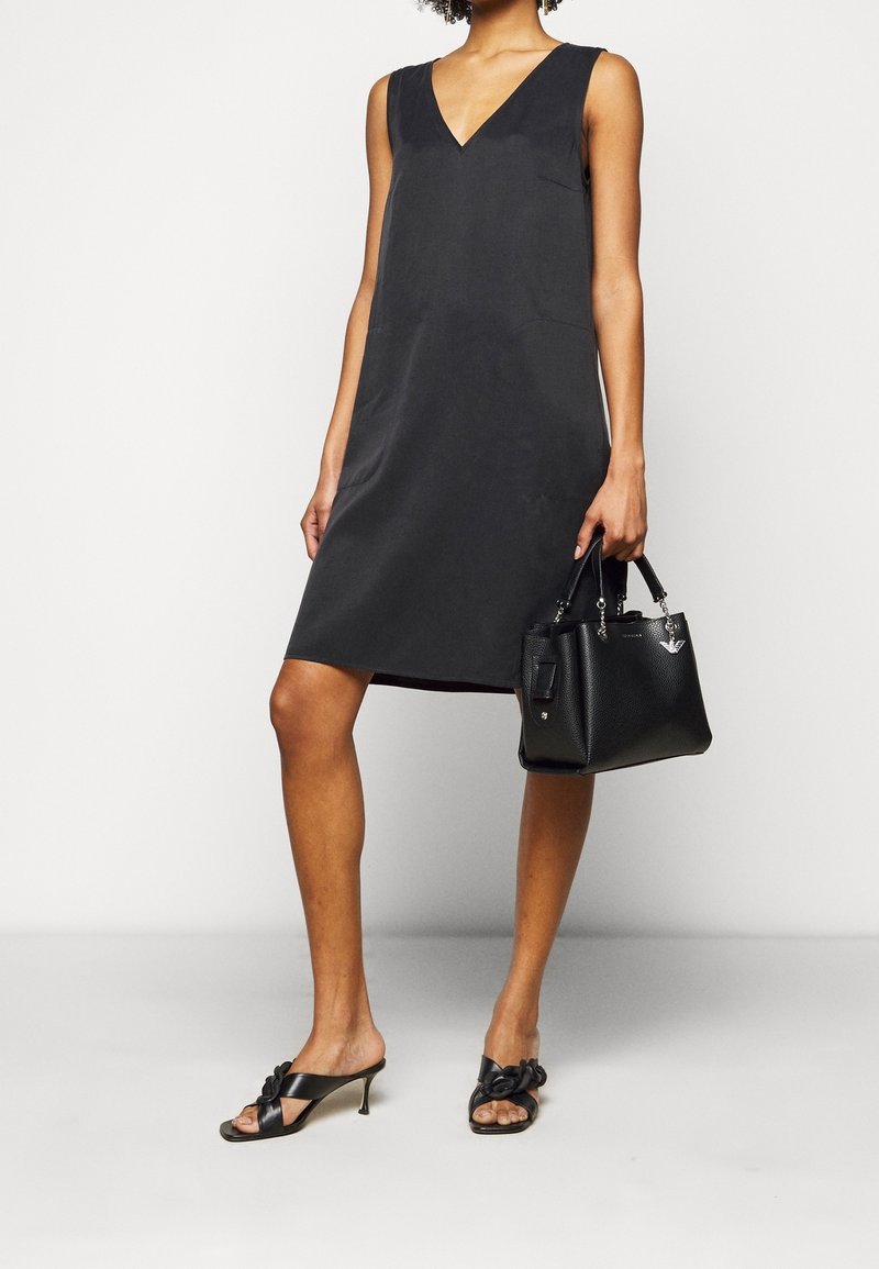 Czarna sukienka bez rękawów z dekoltem w kształcie litery V, gładki materiał, długość do kolan, zestawiona z czarnymi sandałami na obcasie z odkrytymi palcami i czarną, usztywnioną torebką.