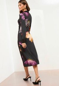 Rochie lungă neagră cu mâneci lungi, cu un model floral multicolor; are o fanta laterală și un design mulat. Accesorizată cu pantofi cu toc negri.