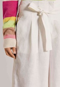Witte linnen broek met wijde pijpen, een strikdetail aan de taille en voorplooien. Een kleurrijke gestreepte mouw van een trui is gedeeltelijk zichtbaar.