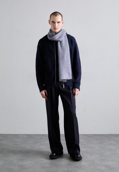 Tamsiai mėlyna fliso džemperis su užtrauktuku, dėvimas ant juodos palaidinės, derinamas su laisvais juodais keliais ir šviesiai pilku šaliku.