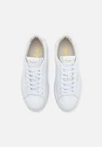 Zapatillas de piel blanca con una textura suave, diseño con cordones y un logo dorado en la lengüeta. La suela es plana y minimalista.