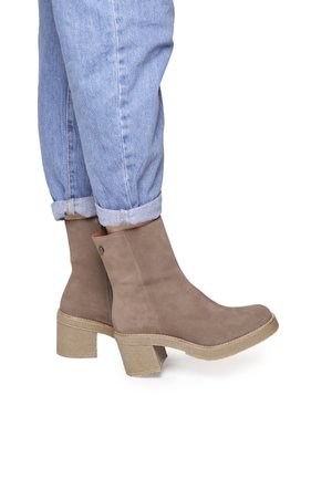 Bottines en daim marron avec une semelle texturée épaisse, dotées d'un bout arrondi et d'un détail avec bouton sur le côté, portées avec un pantalon en denim à un ourlet retroussé.