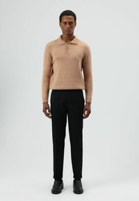 Beige gebreide polotrui met een biezenhals en drie knopen, gecombineerd met zwarte op maat gemaakte broek en zwarte schoenen, tegen een neutrale achtergrond.