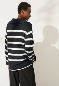 Zwart-wit gestreepte gebreide trui met een brede kraag, lange mouwen en een losse pasvorm, gecombineerd met zwarte broek. Schaduw zichtbaar op de muur.