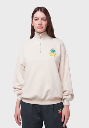 Sweatshirt couleur crème avec un col zippé à quart, des poignets et un ourlet côtelés. Présente une broderie colorée d'un personnage de dessin animé accompagnée de texte.