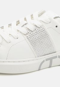 Sneaker in pelle bianca con un pannello di strass argentati texture, lacci bianchi e logo grigio sulla suola. Design classico a ballerina.