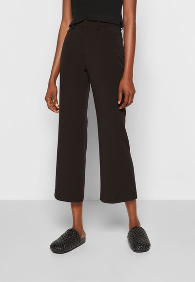 Calças largas pretas feitas de um tecido suave, com uma cintura alta e detalhes de costura subtis. Combinadas com sapatos slip-on pretos em tecido trançado.