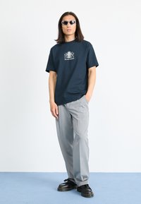 Marineblå crewneck t-skjorte med grafisk trykk, sammen med grå bukser, svarte plattformsko og ovale solbriller. Minimalistisk design.