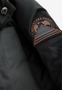 Svart tygjacka med en texturerad finish. Höger ärm har en präglad patch med ett berg- och globmotiv, med orangea accenter.