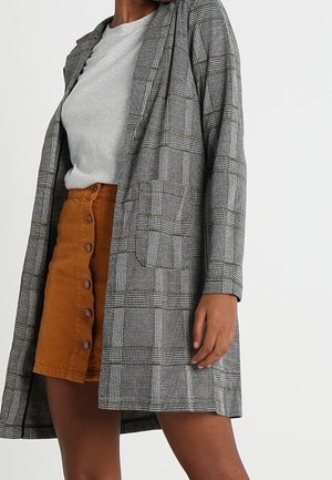 Kobieta w szarym kraciastym długim blezerze, jasnoszarej bluzce i zapinanej na guziki brązowej sztruksowej spódnicy, stojąca z jedną ręką wzdłuż ciała.
