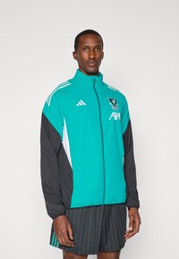 Teal og sort atletisk jakke med hvide accenter, fuld lynlås, stående krave og logo. Båret over sorte stribede shorts.
