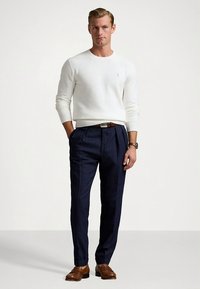 Camisola branca de malha, calças azul-marinho com riscas brancas, cinto de couro castanho e sapatos a condizer. Homem em pé, confiante, mãos ao lado, fundo neutro.