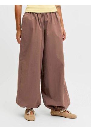 Person iført løstsittende, brune bukser i ballongstil med elastiske mansjetter, kombinert med beige ballettsko, stående mot en ensfarget bakgrunn.