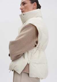 Gepolsterte cremefarbene Weste mit hohem Kragen, die über einer strukturierten beigen Strickjacke getragen wird und eine taillierte Taille sowie seitliche Reißverschlusstaschen aufweist.