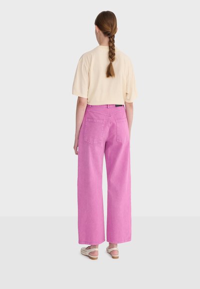 Femme aux cheveux tressés portant un haut crème à manches courtes, un pantalon large rose et des sandales beiges, debout de dos sur un fond uni.