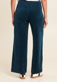 Pantalons en velours côtelé large de couleur teal avec une finition texturée et côtelée. Ils présentent une taille élastique, tombant lâchement au niveau des chevilles, associés à des baskets blanches.
