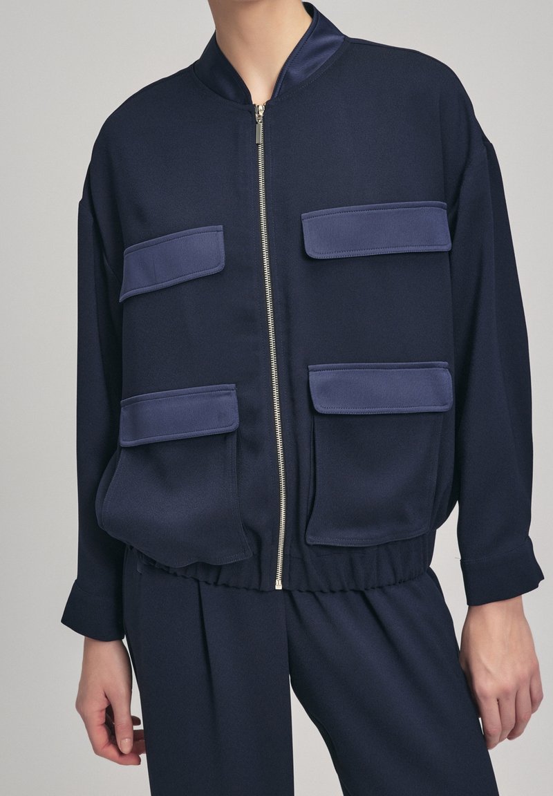 Veste bleu marine zippée avec quatre poches à rabat portée avec un pantalon bleu marine assorti, montrée des épaules jusqu'à mi-cuisse.