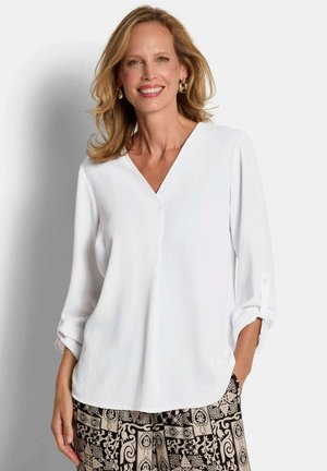 Frau lächelt, trägt eine weiße langärmlige Bluse mit V-Ausschnitt und hochgekrempelten Ärmeln sowie gemusterte schwarze und beigefarbene Hosen.