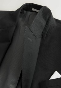 Schwarzer Blazer aus strukturiertem Stoff, mit schlankem Kragen und einem kontrastierenden weißen Einstecktuch. Gefüttertes Innenfutter mit dezente Musterung.