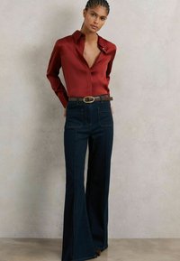 Bluză roșie din satin cu guler și închidere cu buton, asortată cu blugi negri din denim evazați, având detalii vizibile ale cusăturilor și o curea maro.