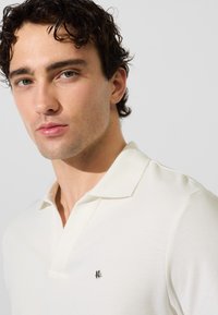 Camiseta de polo blanca con un patrón de tejido texturizado y un cuello clásico, que presenta un pequeño logo metálico en el pecho.