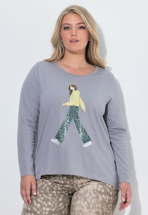 Mujer con camiseta gris de manga larga con un gráfico de una mujer caminando y pantalones beige con estampado sobre un fondo liso y claro.