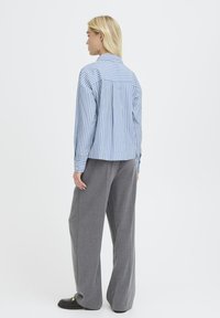 Eine Frau trägt ein blau-weiß gestreiftes Langarmshirt, graue weit geschnittene Hosen und schwarze Slipper. Sie steht mit dem Rücken zu einer weißen Wand.