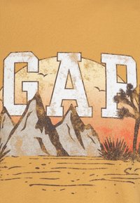 T-shirt graphique jaune avec un grand texte blanc "GAP" au-dessus d'une scène désertique avec des montagnes, des cactus et un fond de coucher de soleil dégradé.