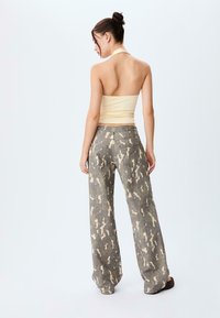 Vrouw met opgestoken haar, gekleed in een crèmekleurige haltertop en loszittende camouflagepatroon broek, staande tegen een eenvoudige witte achtergrond.