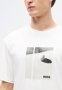 Λευκό βαμβακερό t-shirt με γραφικό σχέδιο μιας μπάλας του τένις και σκιάς. Η μάρκα "BOSS" είναι τυπωμένη στο κάτω μέρος με μαύρο χρώμα.