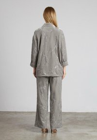 Mujer con el cabello rubio largo viste un traje suelto gris con flores y lentejuelas, con pantalones anchos y mangas remangadas, de pie sobre un suelo pulido.