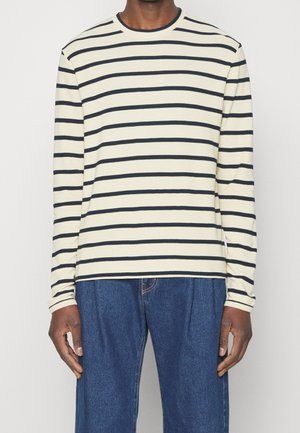 Person trägt cremefarbenes Langarmshirt mit schwarzen horizontalen Streifen und lockere blaue Jeans, steht vor einem schlichten Hintergrund.