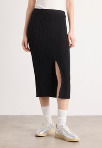 Jupe midi noire en tricot avec des côtes verticales et une fente sur le côté ; associée à des baskets argentées et des chaussettes blanches.