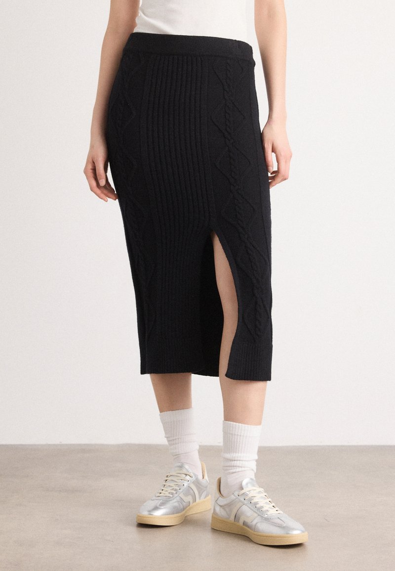 Jupe midi noire en tricot avec des côtes verticales et une fente sur le côté ; associée à des baskets argentées et des chaussettes blanches.