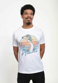 Hvid bomulds T-shirt med et grafisk motiv af et stiliseret dyr og teksten "Hakuna" nedenfor. Billedet har retro, pastelfarvede striber.