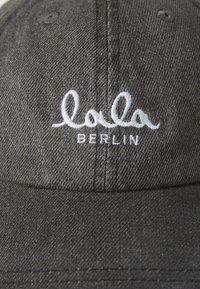 Szürke farmer sapka fehér hímzett "lola BERLIN" felirattal. Strukturált dizájn, ívelt karimával és varrási részletekkel.