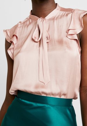 Femme portant un chemisier en satin rose clair avec des manches courtes volantées et un nœud noué au cou, assorti d'une jupe en satin bleu sarcelle.