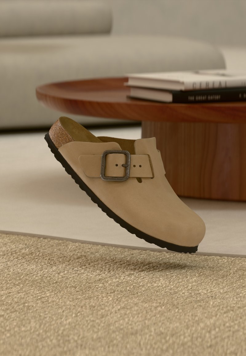 Μπεζ παντόφλα-slip-on με μαύρη σόλα και μεγάλο μεταλλικό πόρπη, τοποθετημένη πάνω σε υφασμένο μπεζ χαλί σε μοντέρνο σαλόνι.