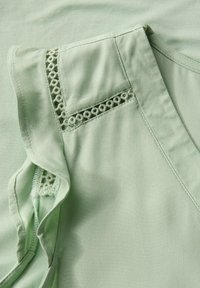Hellgrüne Bluse mit Rüschensäumen und Spitzenbesatz sowie einem runden Halsausschnitt. Weiche Textur mit glattem Stofffinish.