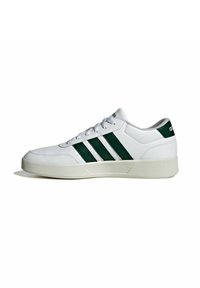 Zapatilla deportiva blanca con textura lisa de cuero, que presenta tres franjas verdes, puntera redonda y suela acolchada.