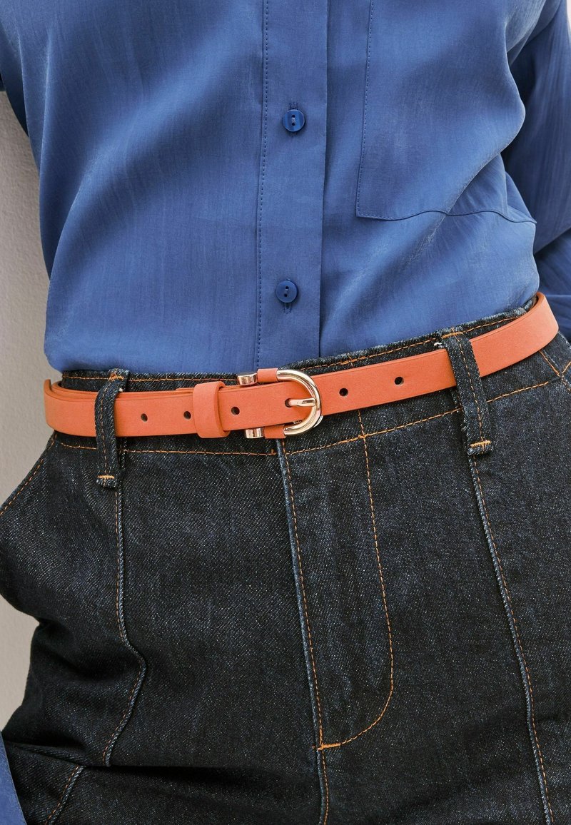 Personne portant une chemise bleue boutonnée rentrée dans un jean en denim foncé avec des surpiqûres orange et une ceinture en cuir orange avec une boucle dorée.