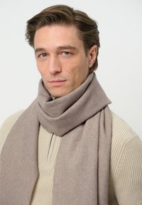 Sciarpa di lana beige avvolta attorno al collo, caratterizzata da una texture morbida e un design semplice. Indossata con un maglione beige chiaro con texture a costine.