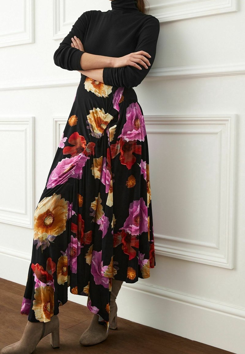 Maxime-kjole med sort bund, prydet med store blomster mønstre i rød, gul og lilla; stoffet falder blødt og er fremhævet af læg.