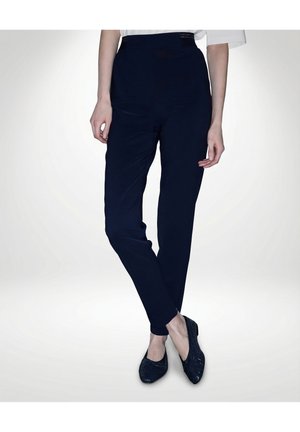 Person trägt eng anliegende marineblaue High-Waist-Hose und schwarze, strukturierte Slipper, steht mit gekreuzten Beinen vor weißem Hintergrund.