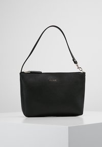 Pochette en cuir noire Guess avec fermeture éclair argentée et bandoulière amovible, exposée sur un socle blanc sur un fond uni.