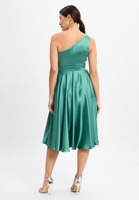 Asymmetrische teal jurk met één schouder in satijnen stof, aansluitende bovenkant, vloeiende rok en glanzende zilveren hoge hakken.