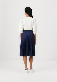 Woman with long braided hair wearing a white puff-sleeve top, navy pleated midi skirt, and white sneakers, standing facing a white wall.