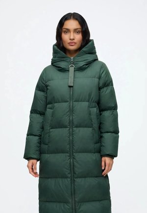 Mujer con un abrigo acolchado largo de color verde oscuro con cuello alto y cremallera frontal, de pie frente a un fondo blanco liso.